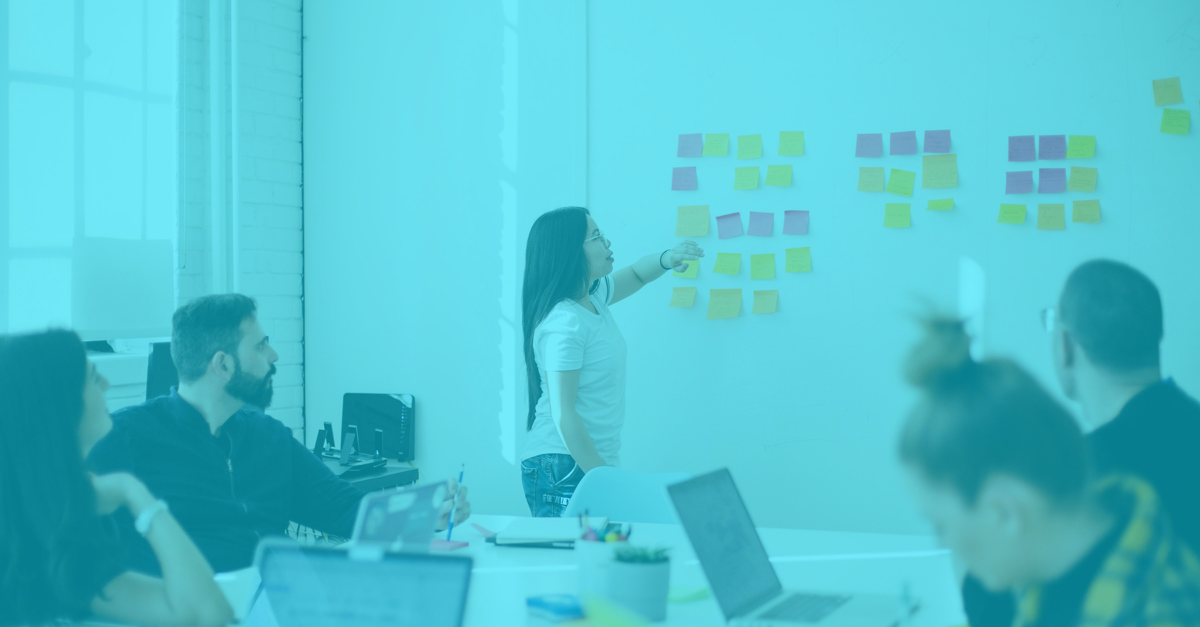 A group sits around a table with laptops open as someone points to post it notes on a whiteboard.