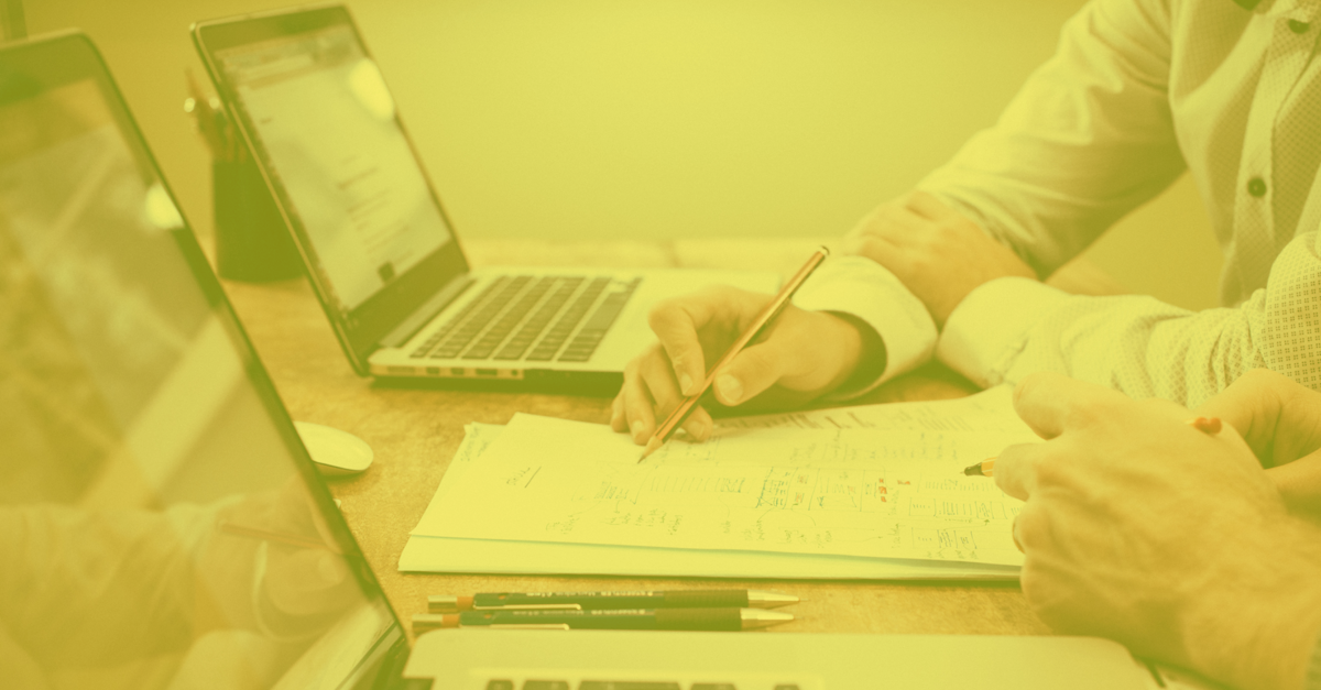 Two laptops open on a conference room table with a mans hand holding a pen writing on a stack of reports